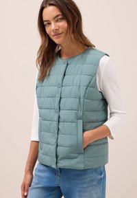 Ljus teal pufferväst med rund halsringning, framtill knäppning med knappar och sidofickor. Bärs över en vit långärmad topp.