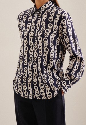 Chemise bleu marine boutonnée avec imprimé de chaîne blanche liée, manches longues, portée rentrée dans un pantalon noir, mains dans les poches.