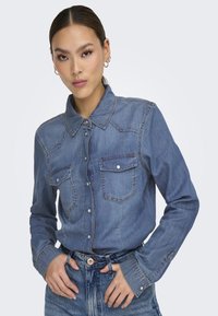 Camisa de mezclilla en azul claro, con cuello puntiagudo, cierre de botones, dos bolsillos en el pecho y mangas largas. Presenta costuras en tono y botones plateados.
