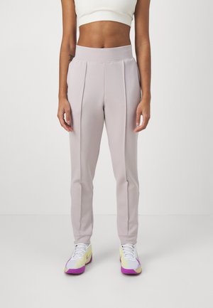 Vrouw draagt lichtpaarse hoge taille broek, wit cropped top en witte sneakers met paarse en gele accenten, staand op een witte vloer.