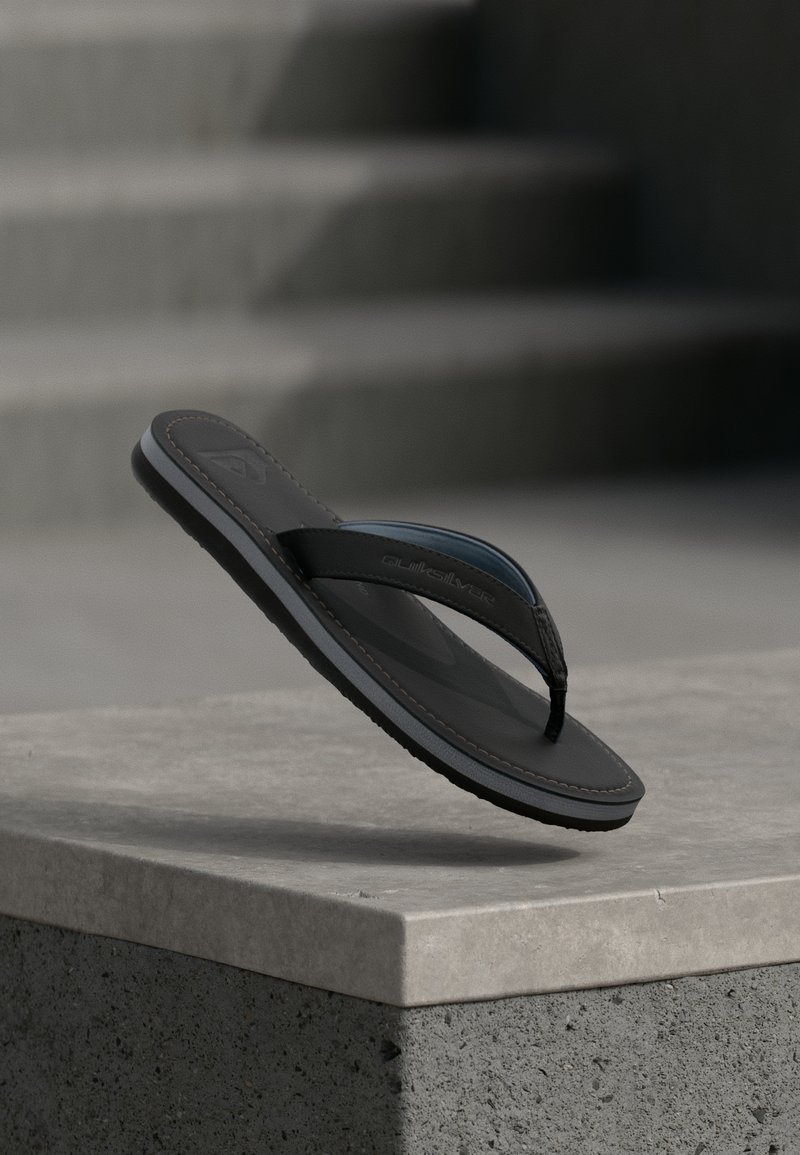 Črna Quiksilver japanka viseča nad betonskim robom z zamegljenimi stopnicami v ozadju.