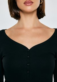 Vrouw die een zwart geribd, geknoopt topje met een brede V-hals draagt, gouden hoepeloorbellen en glanzende roze lippenstift, tegen een effen achtergrond.