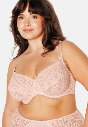 Donna sorridente con capelli castani di media lunghezza che indossa un reggiseno in pizzo rosa chiaro e biancheria intima a vita alta abbinata su sfondo bianco.