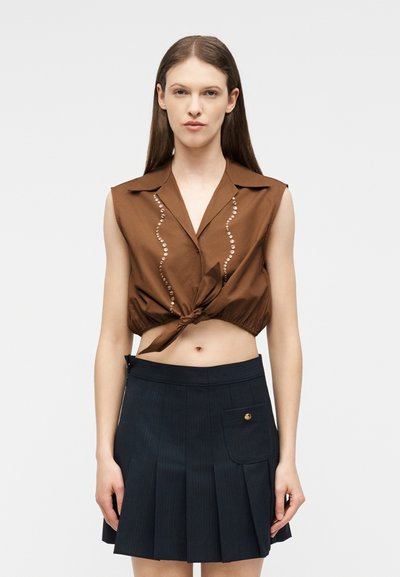 Femme portant un haut marron sans manches, court avec un nœud à l'avant, et une jupe plissée bleu marine avec poche et bouton.