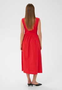 Punane ilma varrukat kleit, millel on sobiv büst ja kogutud seelik, pahkluuni ulatuv, koos mustade terava ninaga muledega. Sile kangastekstuuri.