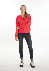 Koronkowy sweter w kolorze koralowym w luźnym kroju, zestawiony z ciemnoniebieskimi dżinsami oraz beżowymi lace-up botkami za kostkę. Gładka faktura i widoczne wzory.