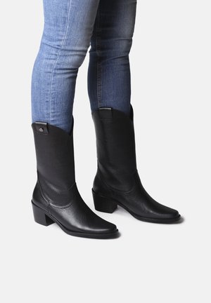 Bottes de cowboy en cuir noir avec un léger talon, bout pointu et détails de couture sur les côtés. Portées avec un jean bleu ajusté.