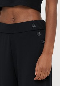 Schwarze Hosen mit weitem Bein sind aus einem glatten Stoff gefertigt, haben einen hohen Bund und dekorative Knopfdetails auf der Vorderseite.