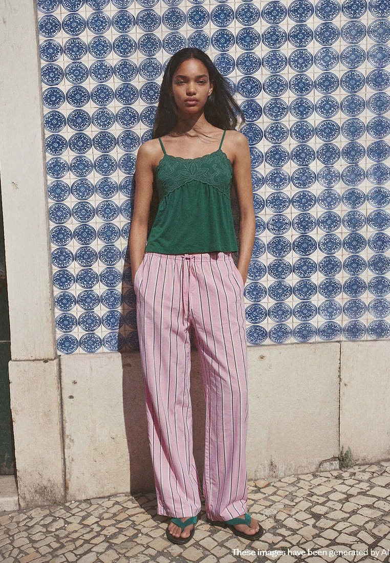 Femme debout contre un mur en carreaux bleu et blanc, portant un haut sans manches vert, un pantalon rayé rose et des sandales vertes, les mains dans les poches.