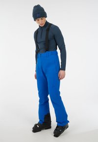 Blauwe skibroek met verstelbare bretels, met een gladde textuur en zwarte accenten bij de enkels; gecombineerd met een marineblauw langemouwen shirt en een beanie.