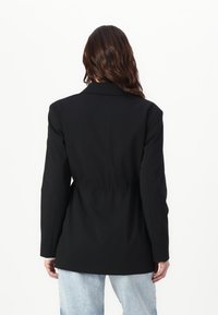 Črn blazer s po meri zasnovano krojenjem, dolgimi rokavi in poudarjenim pasom. Gladka tekstura, brez vidnih vzorcev in standarden ovratnik.