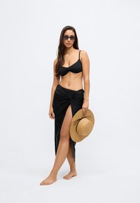 Crni bikini top i suknja s visokim prorezom; drži slamnik s crnom mašnom. Glatka tekstura, pripijeni kroj, minimalistički stil.