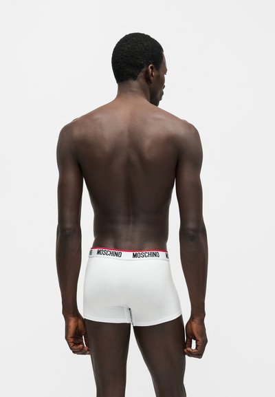 Άνδρας με σκουρόχρωμο δέρμα φορώντας λευκά boxer Moschino, στέκεται με την πλάτη προς το φωτεινό μονόχρωμο φόντο.