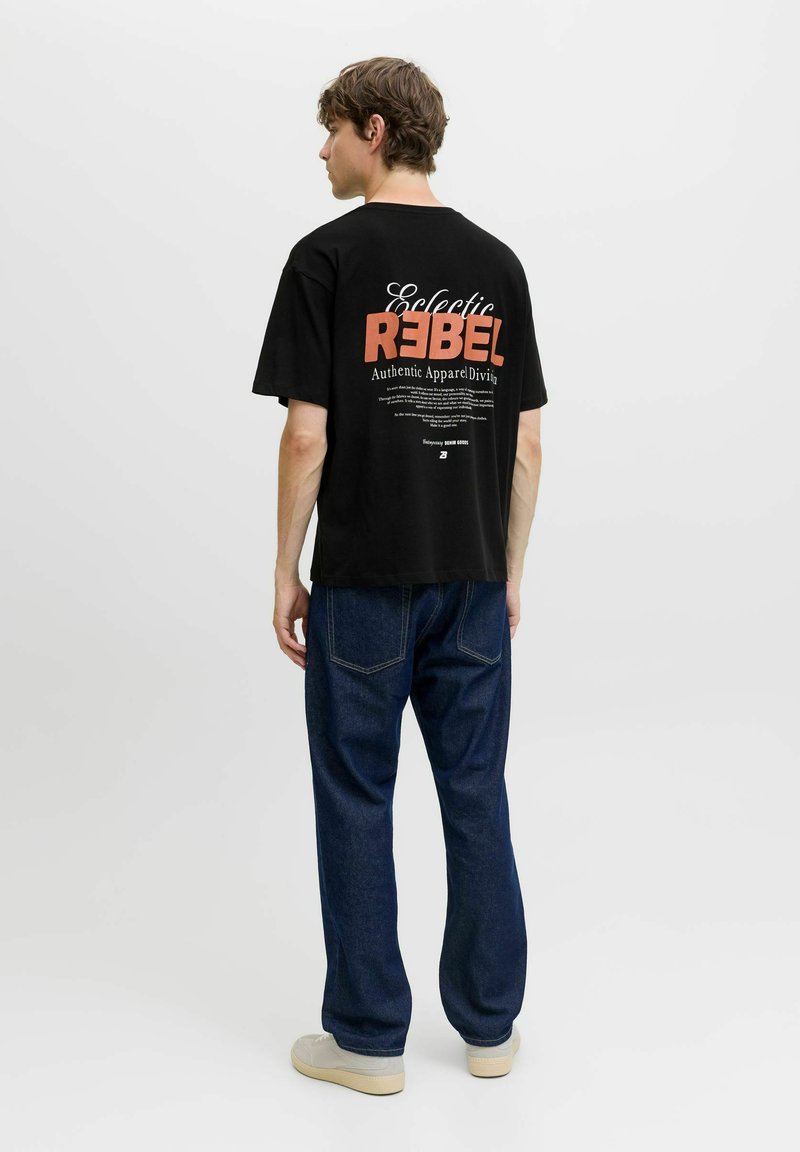 Young man in black graphic T-shirt reading "Eclectic Rebel," blue jeans, and white sneakers standing against plain white background, facing away.