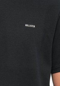 Juoda trumpomis rankovėmis marškinėliai su maža balta "HOLLISTER" tekstu viršutinėje krūtinės dalyje, dėvinčio asmens matomu ranka.