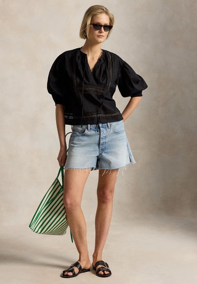 Νεαρή γυναίκα που φοράει μαύρο μπλούζα με φουσκωτά μανίκια, ανοιχτόχρωμο τζιν σορτς, μαύρα σανδάλια, γυαλιά ηλίου, κρατώντας μια τσάντα tote με πράσινες και λευκές ρίγες.