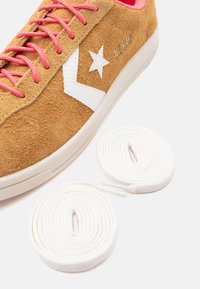 Τακούνια από σαμουά δέρμα με λευκό λογότυπο αστέρι, ροζ κορδόνια και δύο τυλιγμένα λευκά κορδόνια σε λευκό φόντο.