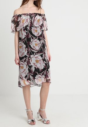Femme portant une robe noire mi-longue à épaules dénudées avec un grand imprimé floral rose et des sandales argentées à talons hauts, debout sur fond blanc.