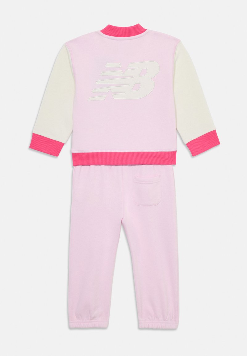 Ensemble de survêtement rose et blanc comprenant une veste zippée avec des poignets et un ourlet côtelés, un logo au dos, ainsi qu'un pantalon assorti avec une taille élastique et une poche.