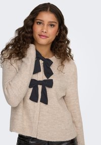 Beige neulepusero, jossa on pitkät hihat ja kaksi mustaa nauhabussia kaula-aukossa, rento istuvuus. Sileä pinta.