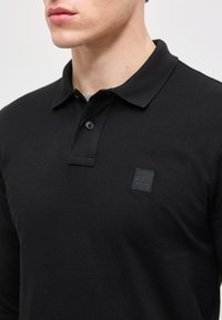 Man draagt een zwart poloshirt met kraag, voorzien van twee knopen en een klein gegraveerd "BOSS"-logo op de borst.