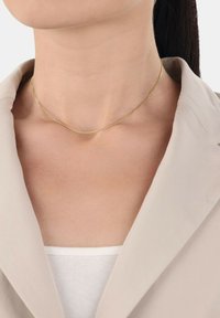 Collana d'oro con un design sottile e delicato, caratterizzata da una superficie liscia e da una leggera brillantezza, indossata su uno sfondo di abbigliamento beige chiaro.