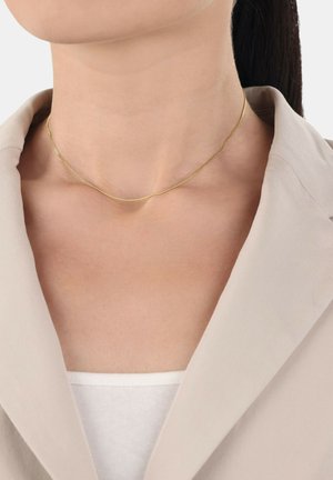 Collana d'oro con un design sottile e delicato, caratterizzata da una superficie liscia e da una leggera brillantezza, indossata su uno sfondo di abbigliamento beige chiaro.