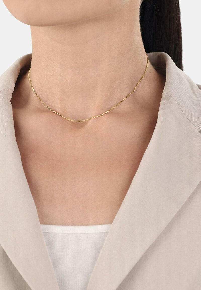 Collana d'oro con un design sottile e delicato, caratterizzata da una superficie liscia e da una leggera brillantezza, indossata su uno sfondo di abbigliamento beige chiaro.