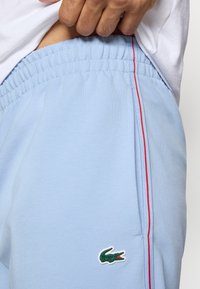 Ανοιχτό μπλε φόρμες με ελαστική μέση, πλαϊνές ρίγες σε κόκκινο χρώμα κα ι πράσινο λογότυπο Lacoste στο αριστερό πλάι.