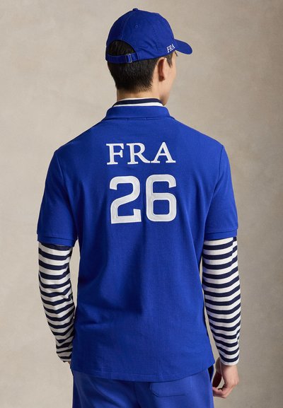 Hombre con gorra azul y polo con "FRA" y número "26" en la espalda, con mangas largas a rayas blancas y negras debajo.