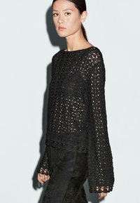 Schwarzer Strickpullover mit offenem Gewebe und ausgestellten langen Ärmeln. Mit gewelltem Saum und strukturiertem Stoff. Kombiniert mit dunklen Wildlederhosen.