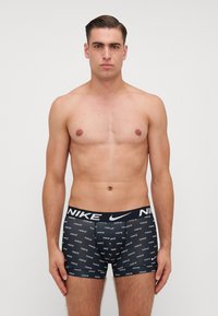 Boxeri negri cu logo alb "NIKE" și un model de text repetitiv, talie elastică și finisaj neted. Modelul stă în picioare.