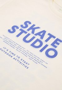 Balta medvilninė marškinė su ryškiai mėlynu tekstu, kuriame rašoma "SKATE STUDIO" ir papildomu tekstu. Minkšta tekstūra ir standartinio apvalaus kaklo dizainas.