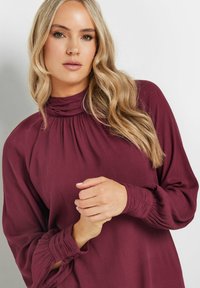 Blusa bordeaux con collo alto arricciato, maniche lunghe e polsini abbottonati. Il tessuto ha una finitura morbida e texture.