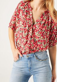 Red and cream floral-patterned blouse with short sleeves, v-neck, and buttons; worn with light blue denim jeans with decorative accents.