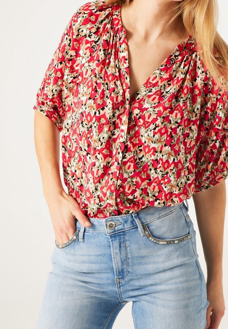 Red and cream floral-patterned blouse with short sleeves, v-neck, and buttons; worn with light blue denim jeans with decorative accents.