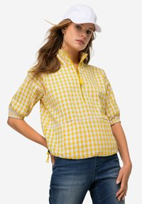 Camicia gialla e bianca a scacchi con zip e maniche corte, in tessuto leggero, dotata di tasca. Abbinata a jeans blu scuro e un cappellino bianco.