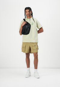 Light green short-sleeve t-shirt with a pocket logo, paired with tan cargo shorts featuring front pockets and a black crossbody bag. White sneakers.