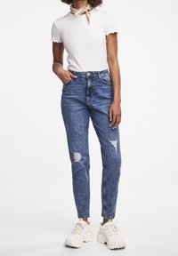 Jeans high-waisted blu chiaro in denim con dettagli strappati, abbinati a una maglietta bianca aderente a coste e sneaker bianche chunky.