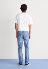 Jeans de mezclilla azul claro con un corte recto, con bolsillos traseros y un parche de cuero con el logo. Combinados con una camiseta de algodón blanca y zapatos negros.