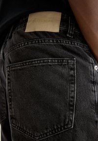 Svarta denimjeans med en läderetikett där "PULL&BEAR" är präglat, synlig steg i bakfickorna och en strukturerad tygfinish.