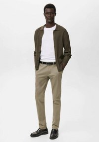 Mörkgrön ribbad cardigan över en vit t-shirt, parat med beige byxor och svarta skor. Enkelt design med knappdetaljer.