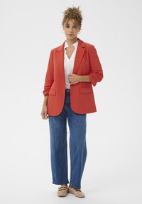 Blazer rouge avec revers crantés, deux poches avant et manches retroussées, associé à un jean droit bleu clair et des chaussures beiges.