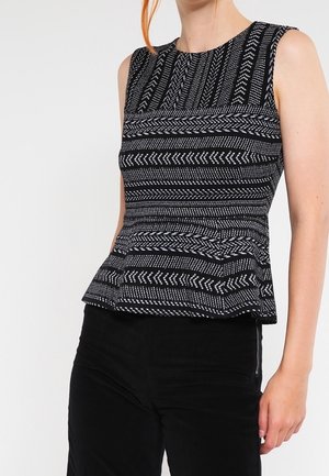 Kobieta w czarno-białym, wzorzystym, bezrękawnikowym topie peplum i czarnych spodniach, stoi na białym tle.