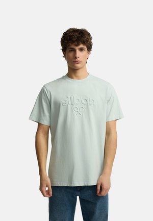 Joven con pelo rizado que lleva una camiseta verde pálido con texto y símbolo "silbon" en relieve, combinada con vaqueros azules, mirando hacia adelante.