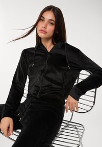 Felpa nera con zip realizzata in morbido materiale velour, dotata di cappuccio, cordini e maniche lunghe, abbinata a pantaloni neri coordinati.