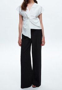 Blusa bianca avvolgente con maniche corte e dettaglio anteriore attorcigliato, abbinata a pantaloni neri larghi. Tessuto liscio con un design pulito.