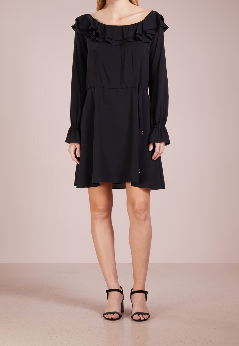 Femme portant une robe noire à manches longues avec un col à volants, une taille cintrée, et des sandales à talons hauts noires à bout ouvert, debout sur un sol beige.