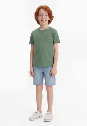 Χαμογελαστό αγόρι με κόκκινα μαλλιά που στέκεται, φοράει πράσινο κοντομάνικο μπλουζάκι, ανοιχτό μπλε τζιν σορτς και λευκά αθλητικά παπούτσια με λευκό φόντο.