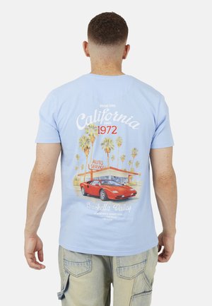 Man met een lichtblauw T-shirt met een vintage California-afbeelding van een rode sportwagen, palmbomen en een autoservicewinkel op de achterkant.